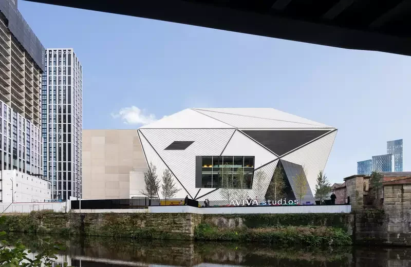 Exterior view of Aviva Studios across the River Irwell, showing the geometric silver building and surrounding city architecture.